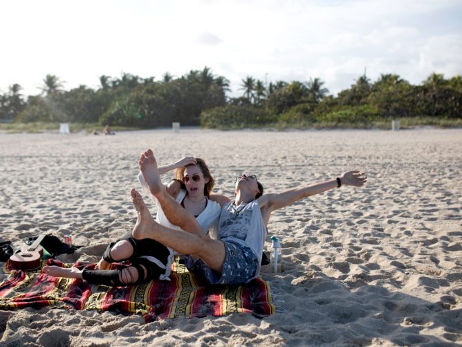Mary Aleda and I laughing and having a good time on the beach with some friends, showing how goofy we are and how much we really love each other. I am wearing a tank top and trunks and she is wearing a white shirt and black skinny jeans sitting on a quilt blanket